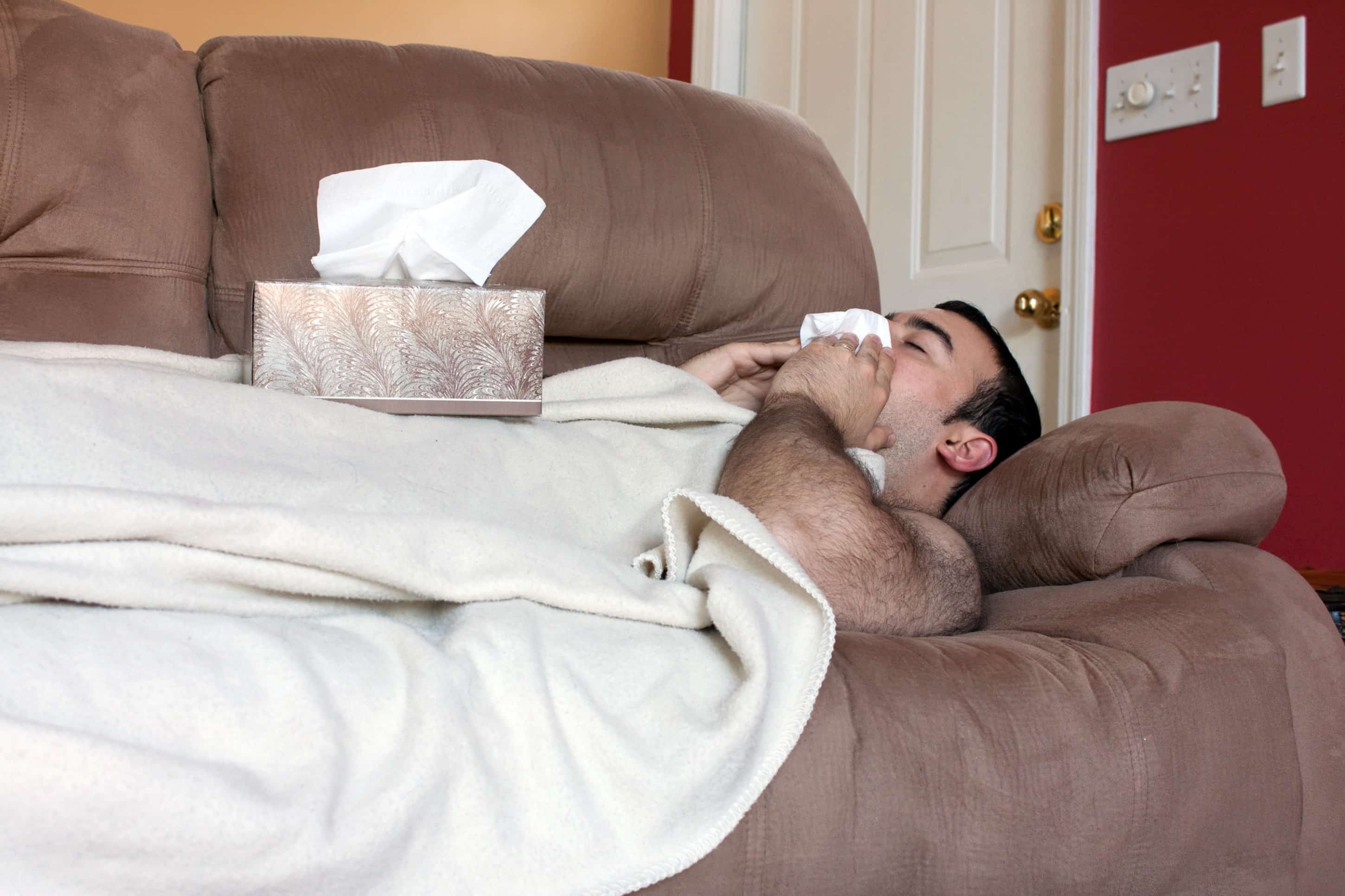 A young adult sick on the couch at home blows his nose with a tissue